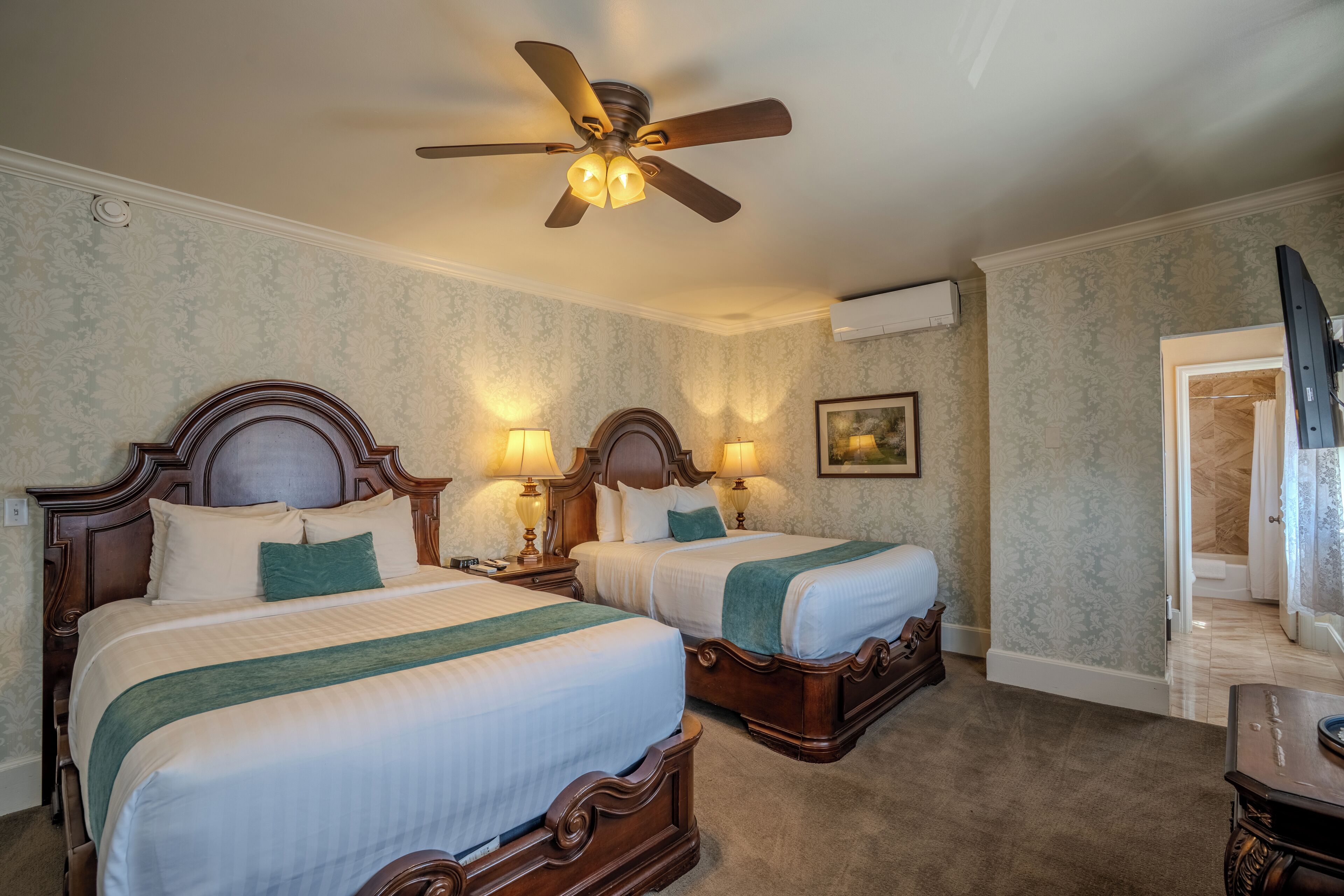 Quarto de hotel com duas camas, um ventilador de teto, luminárias fixadas na parede, um quadro e uma porta de acesso a um banheiro.
