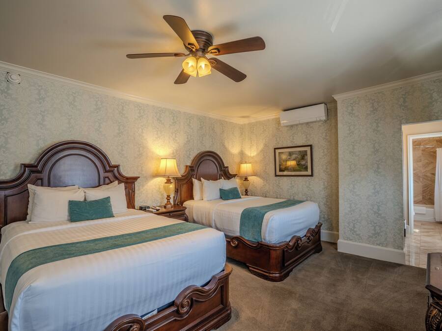 Quarto de hotel com duas camas, um ventilador de teto, luminárias fixadas na parede, um quadro e uma porta de acesso a um banheiro.