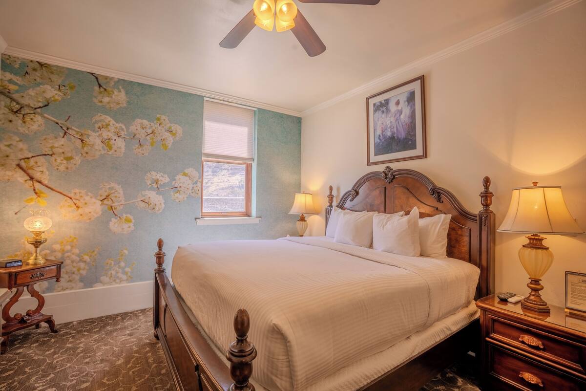 A bedroom with a large bed, two bedside tables with lamps, a ceiling fan, a window with blinds, and a wall with a floral mural.