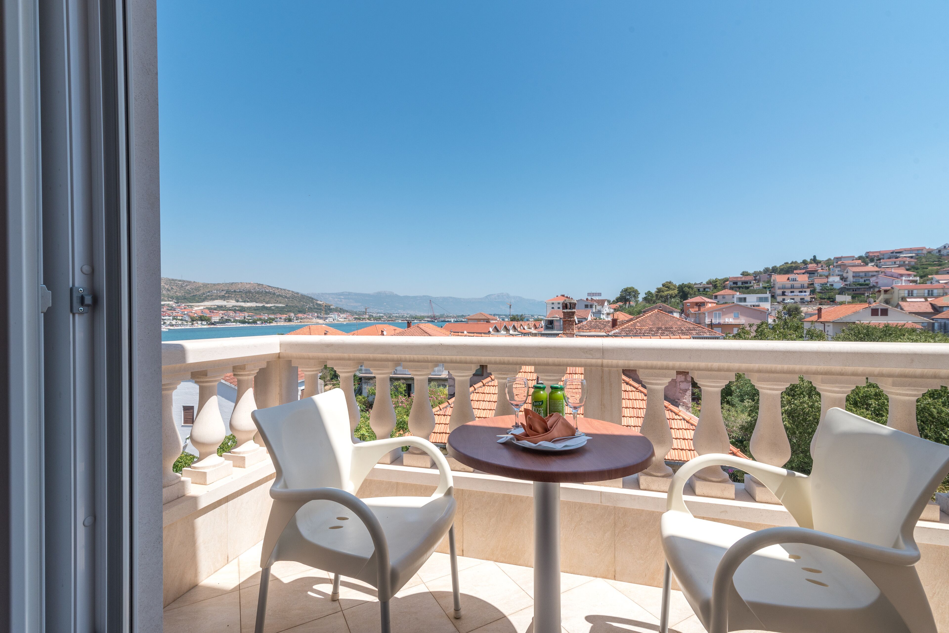 Un balcon avec deux chaises blanches et une petite table ronde, donnant sur une ville côtière aux bâtiments aux toits rouges et sous un ciel bleu dégagé.