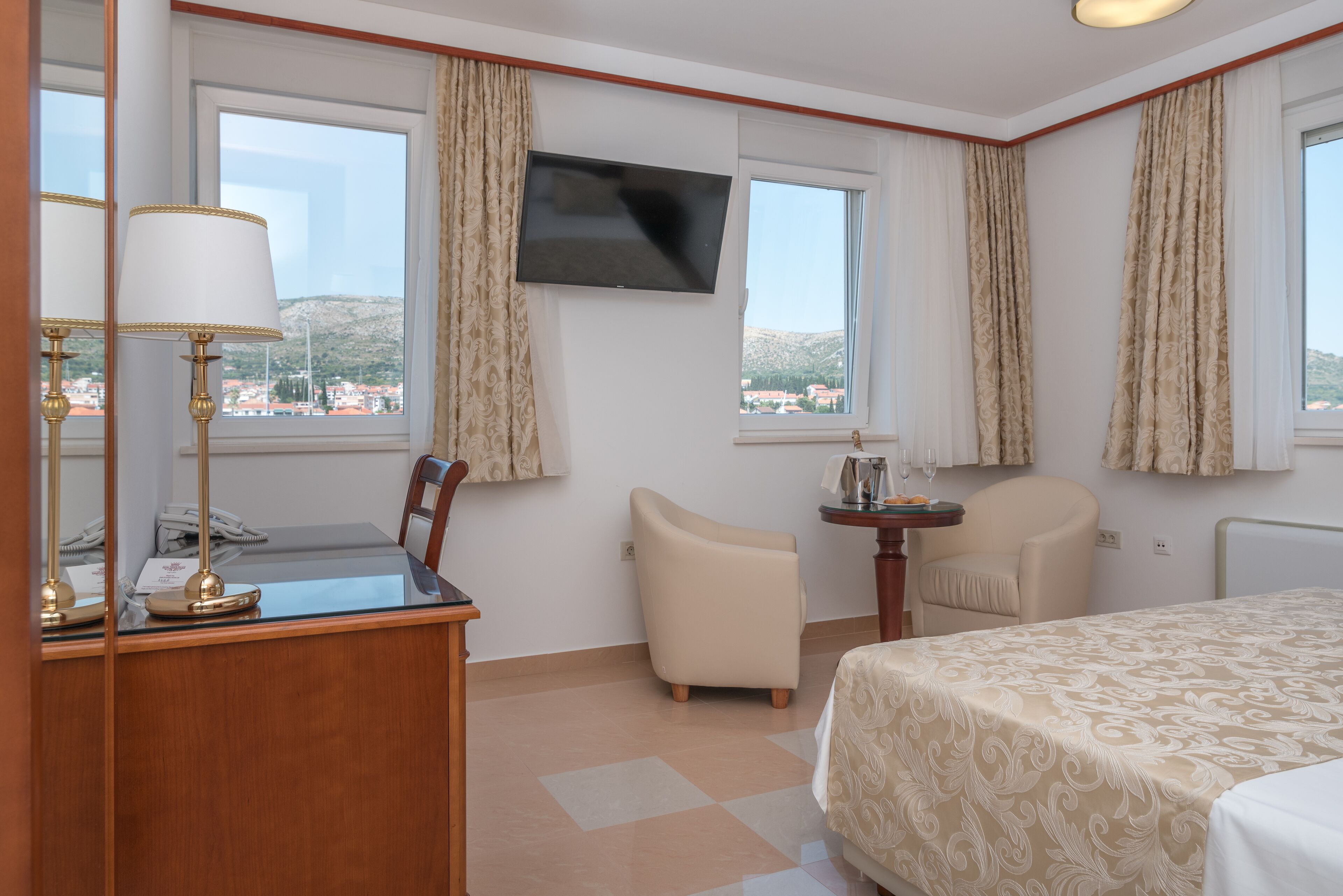 Quarto de hotel com escrivaninha, cadeira, cama, televisão e janelas com cortinas.