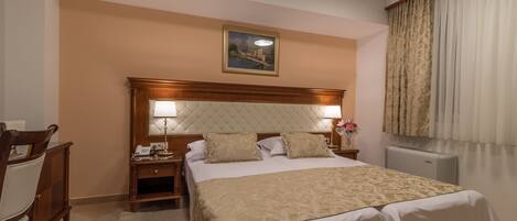 A hotel room with a large bed, a wooden headboard, a nightstand with a lamp, a desk with a chair, and a window with curtains.