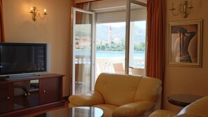 A hotel room with a television, a sofa, a small table, and a balcony with a view of a body of water and buildings. - Hotel Trogir Palace (Trogir)