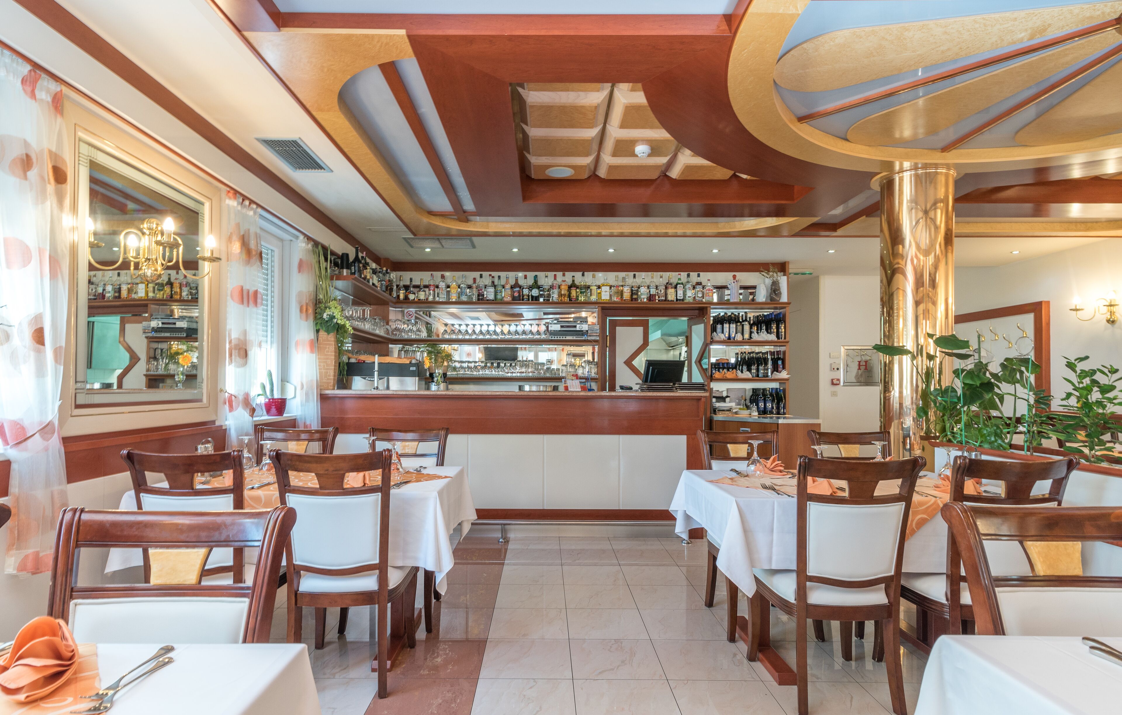 A restaurant interior with wooden tables and chairs, a bar area with shelves of bottles, and a decorative ceiling.