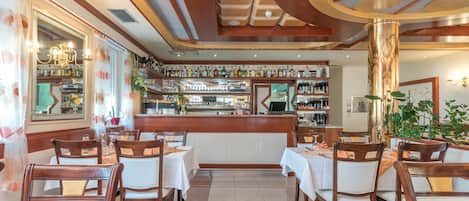A restaurant interior with wooden tables and chairs, a bar area with shelves of bottles, and a decorative ceiling.