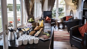 Um buffet de café da manhã com diversos alimentos e bebidas, uma televisão e opções de assento em um ambiente com grandes janelas e vista para o exterior.
