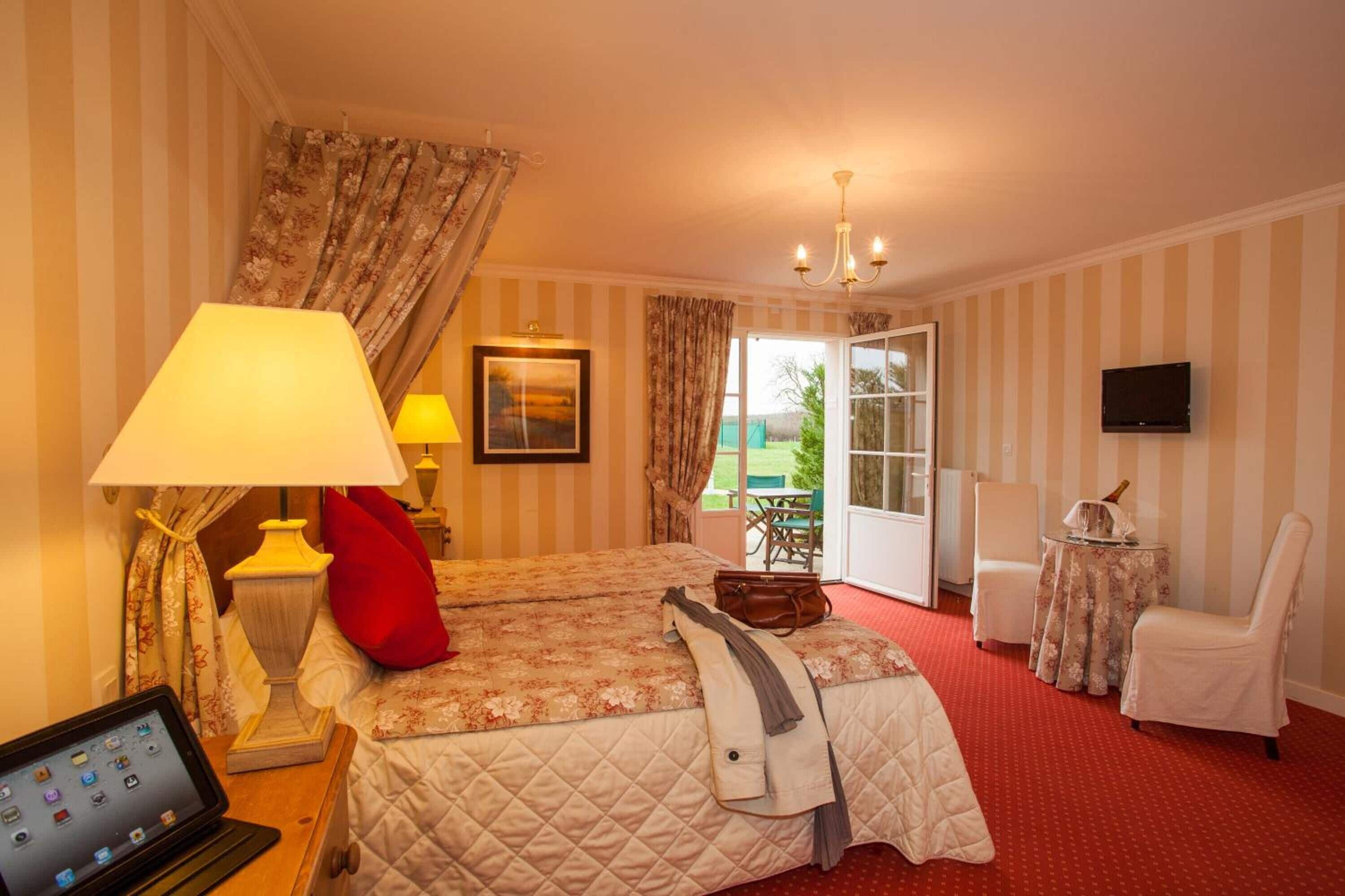 A hotel room with a bed, two bedside lamps, a laptop, a TV, a chair, and a small table with a bottle.