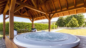Uma banheira de hidromassagem sob uma pérgola de madeira, com vista para um arbusto e uma casa ao fundo.