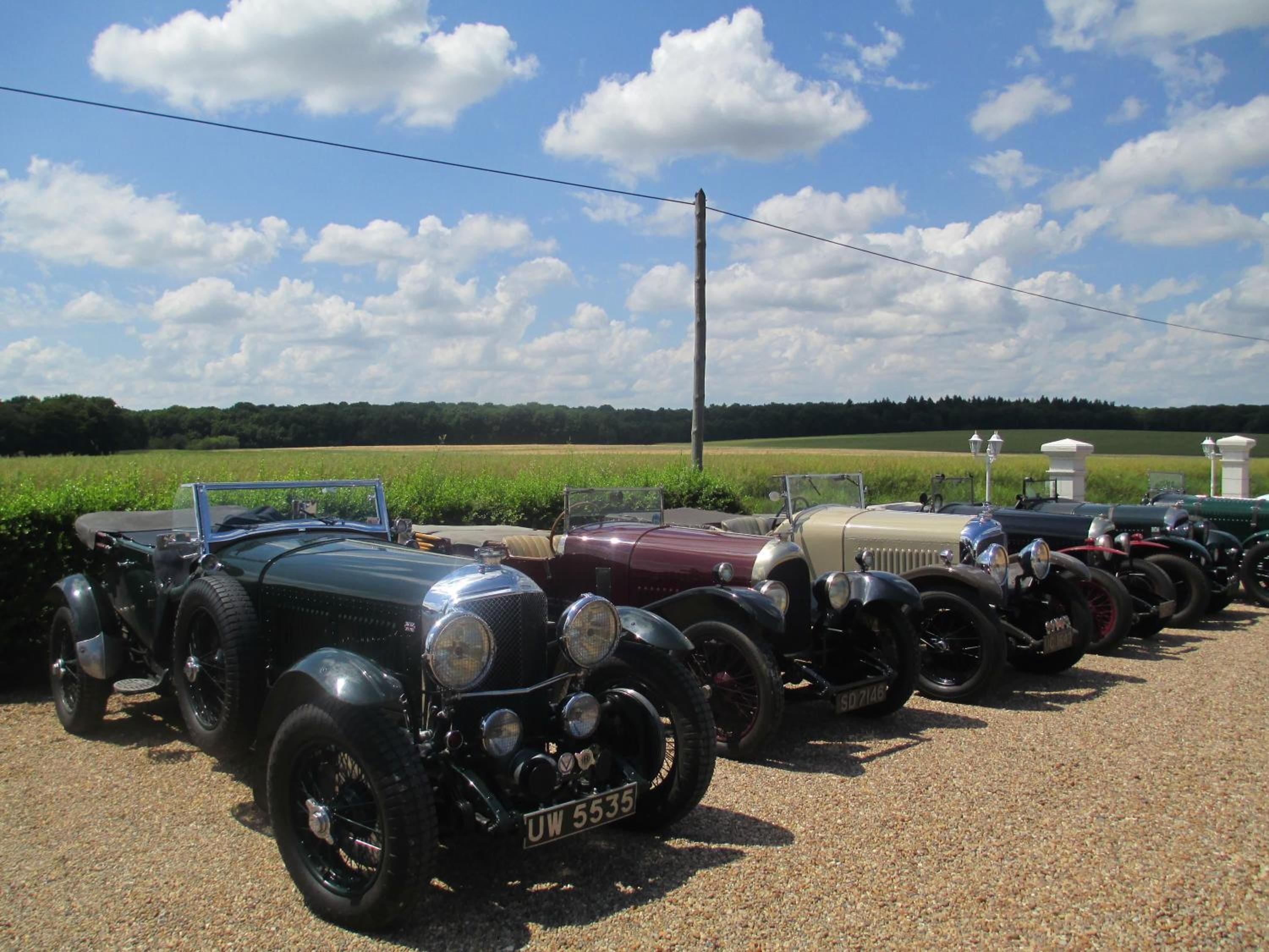 A row of vintage cars parked on a gravel surface with a clear sky and green fields in the background.