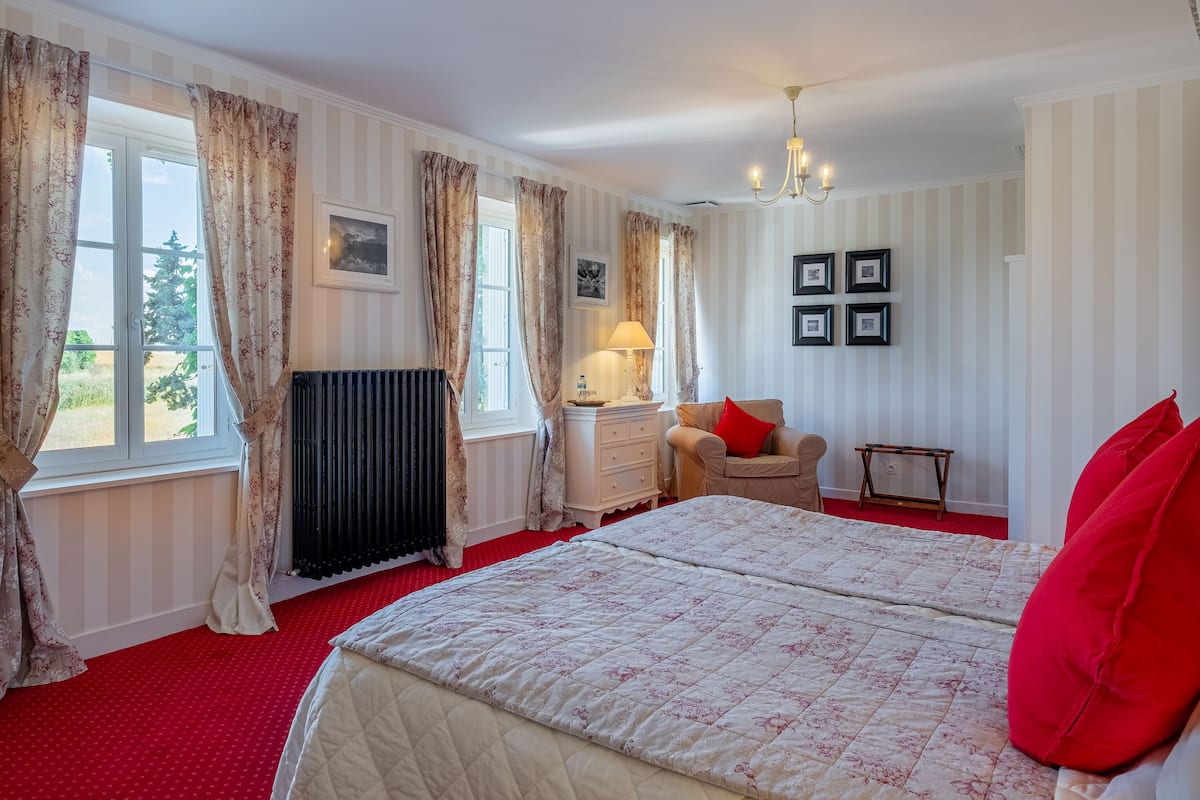 Habitación de hotel con una cama grande, una alfombra roja, un radiador, una mesita de noche con una lámpara, una silla y vistas a zonas verdes.