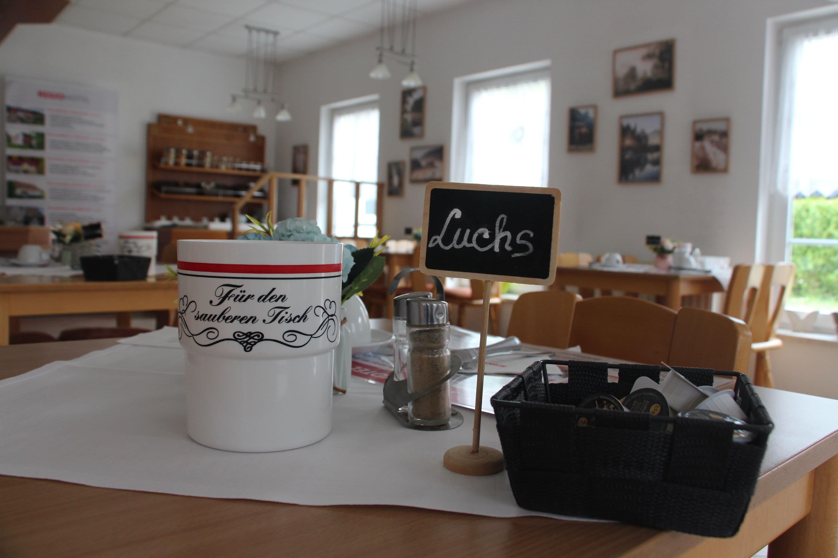 A table with a white mug, a small plant, a salt shaker, and a black basket. A chalkboard sign with the word "Lucks" is also visible.