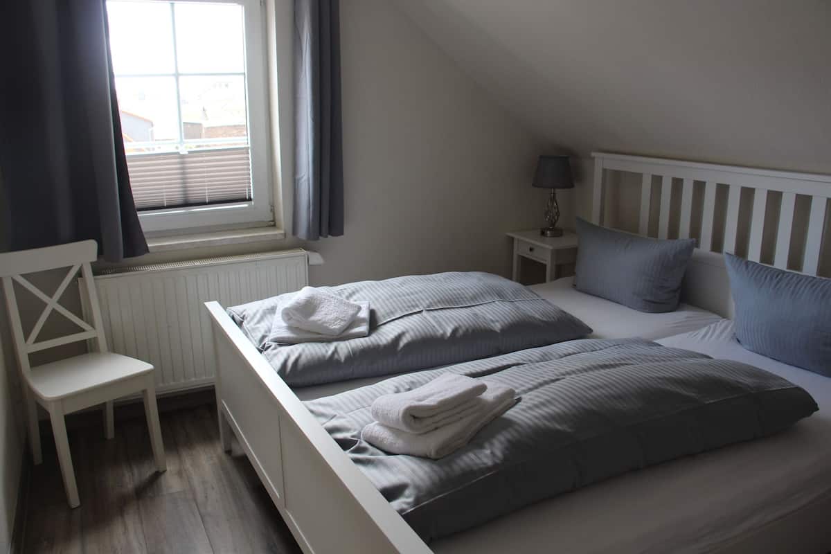 A bedroom with two beds, a chair, a nightstand with a lamp, and a window with curtains.