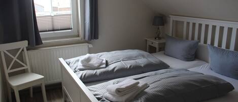 A bedroom with two beds, a chair, a nightstand with a lamp, and a window with curtains.