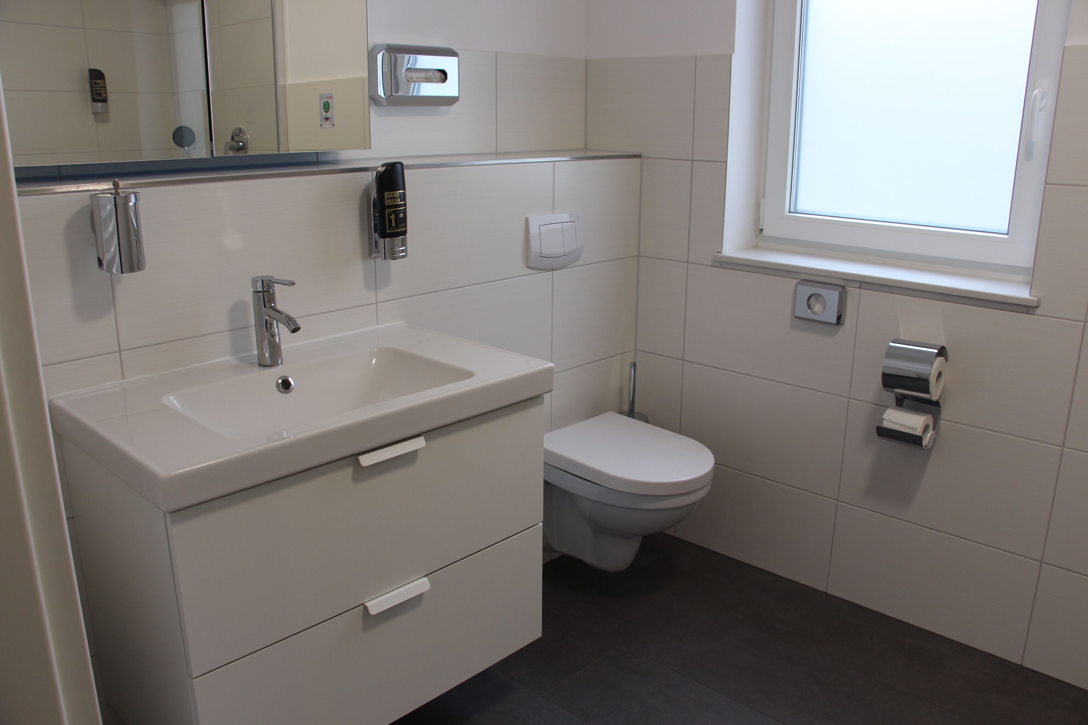 Un baño con lavamanos blanco, inodoro, espejo y ventana.