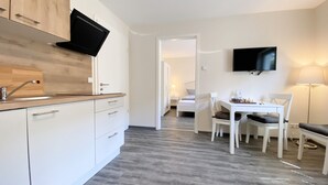 Une cuisine moderne dotée d’un téléviseur encastré, un coin repas avec un jeu d’échecs et une chambre à coucher visible à travers une porte ouverte.