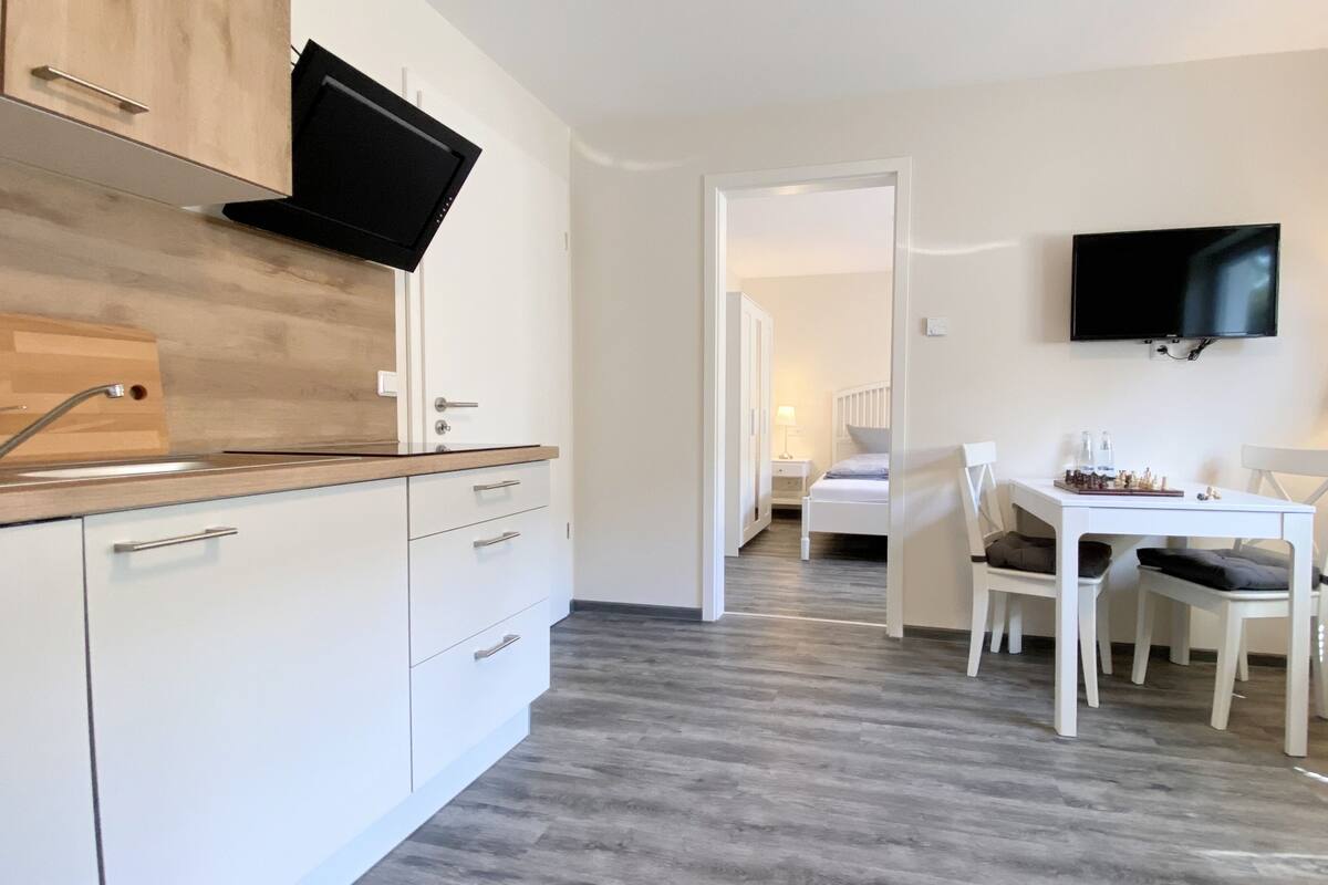 A modern kitchen with a flat-screen TV, a dining area with a chessboard, and a bedroom with a bed and nightstand.