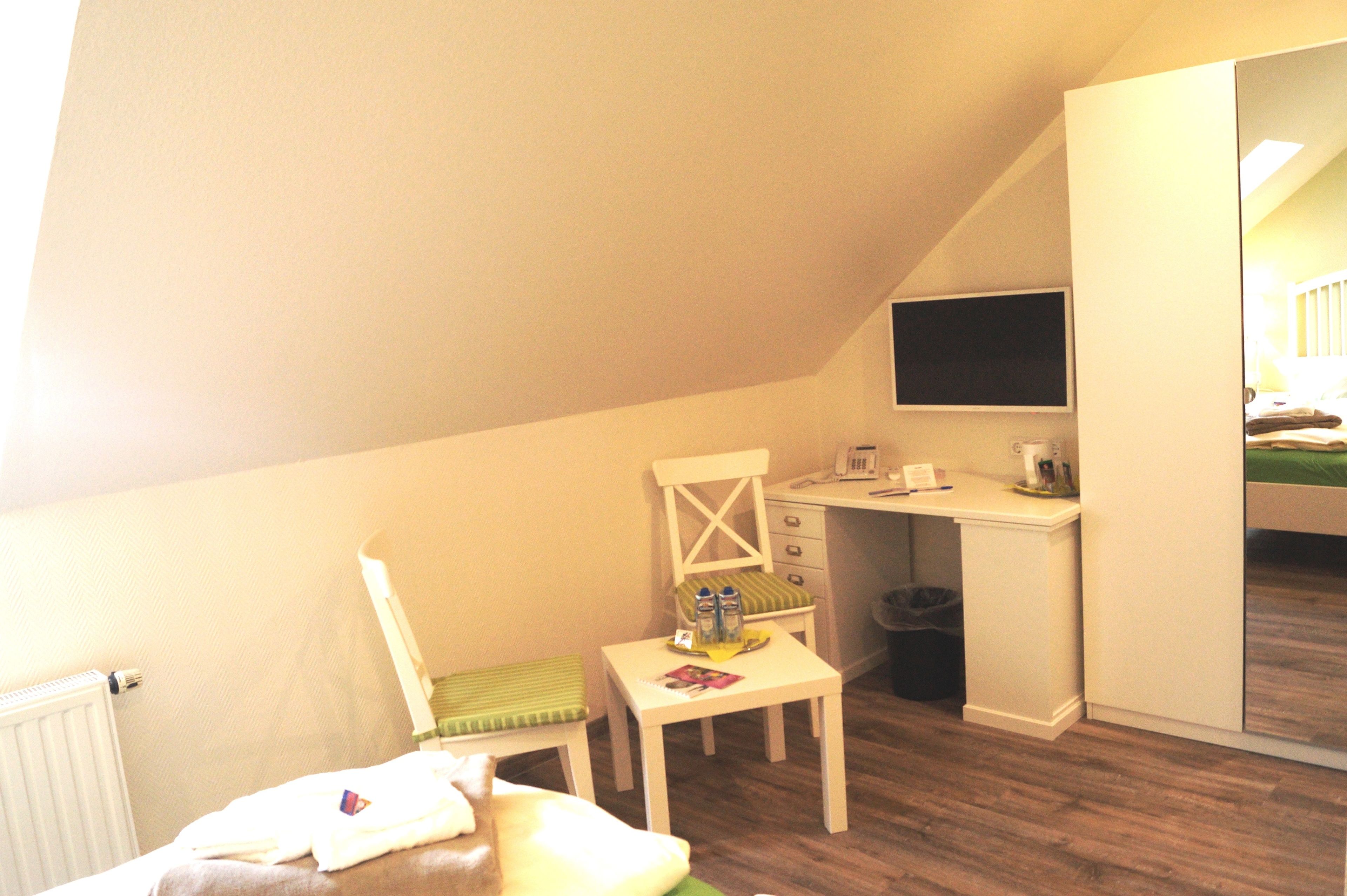 A small, well-lit room with a sloped ceiling, a desk with a chair, a bed, and a television mounted on the wall.