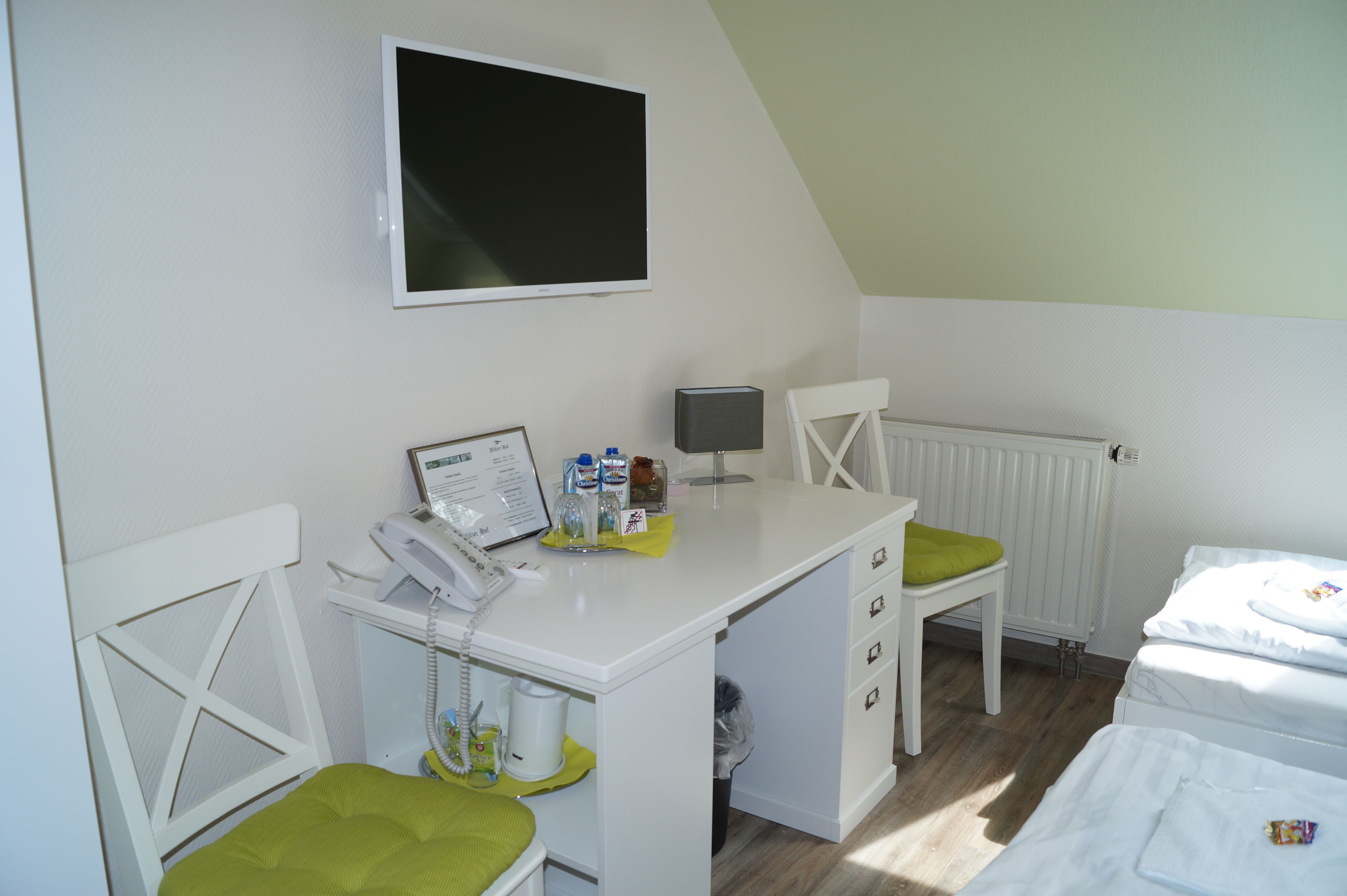 Une petite pièce propre, équipée d’un bureau, d’une chaise et d’un lit. Un téléviseur est fixé au mur, il y a aussi un radiateur et une fenêtre.