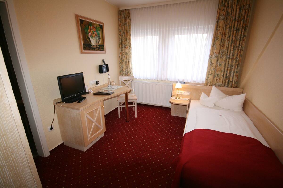 A hotel room with a bed, a desk with a computer, a chair, a window with curtains, and a radiator.