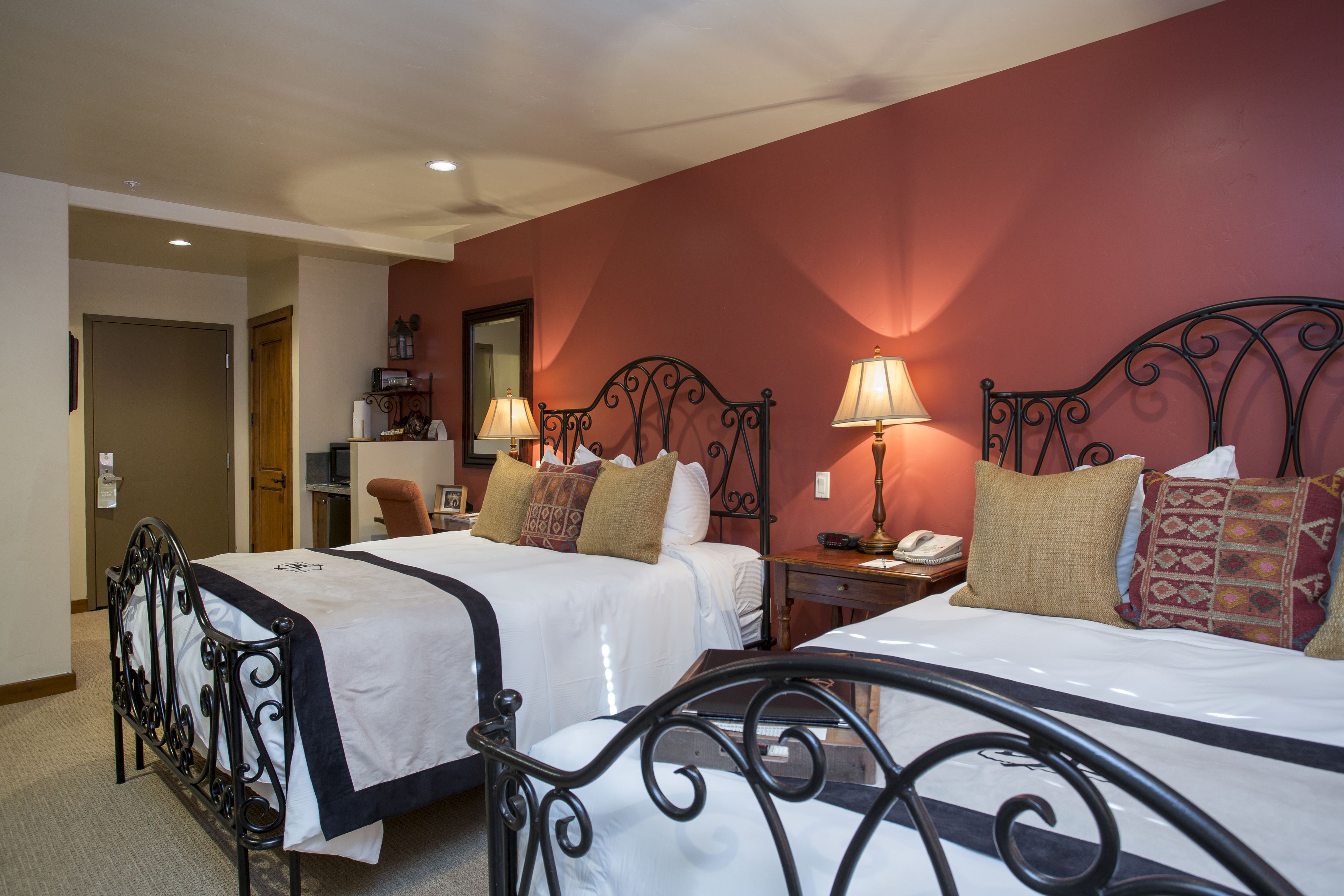 A hotel room with two beds, each with a black metal frame and white bedding. There are two bedside tables with lamps, and a wooden door leading to another room.