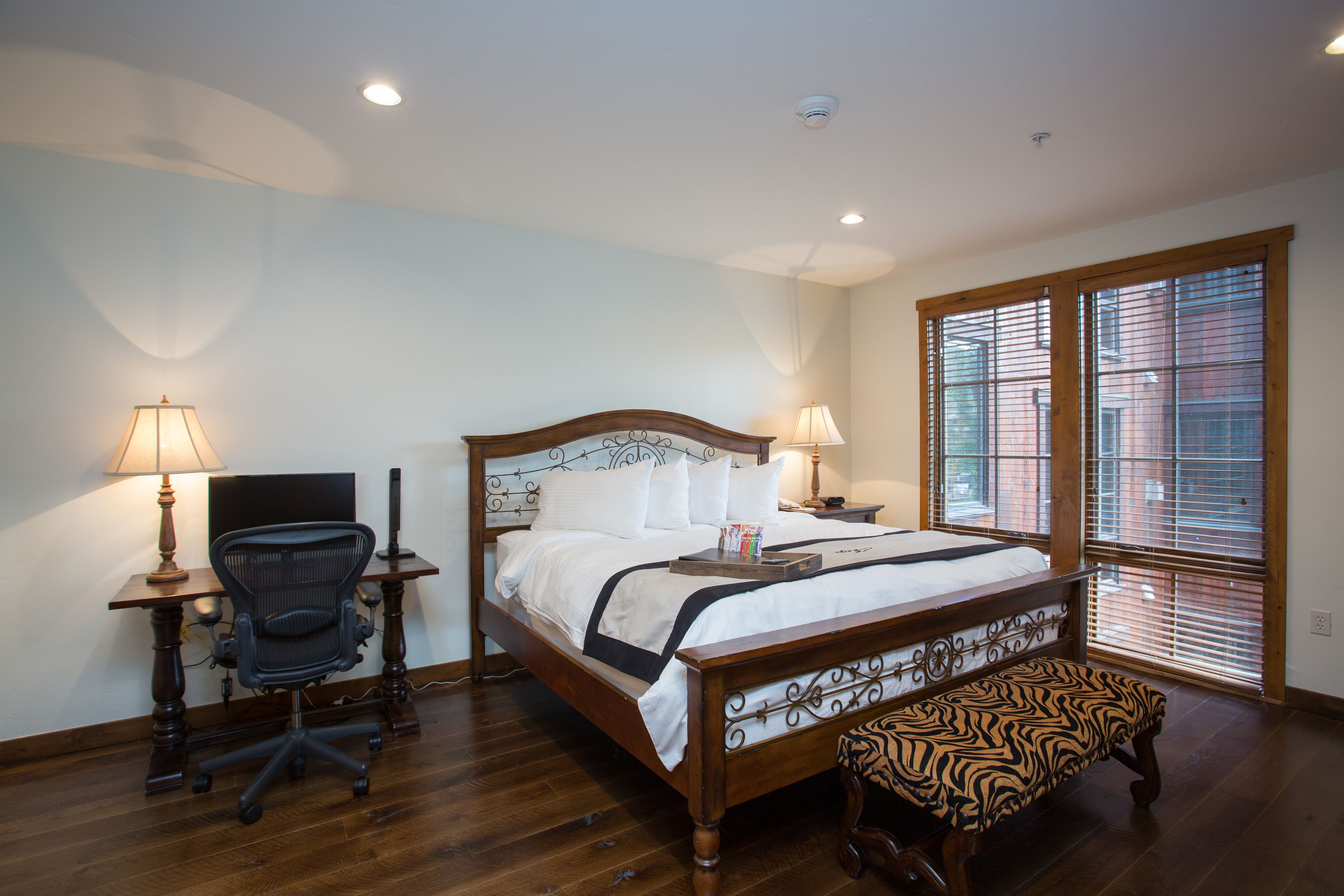 A bedroom with a wooden bed, a desk with a chair, a bench, and a window with blinds.