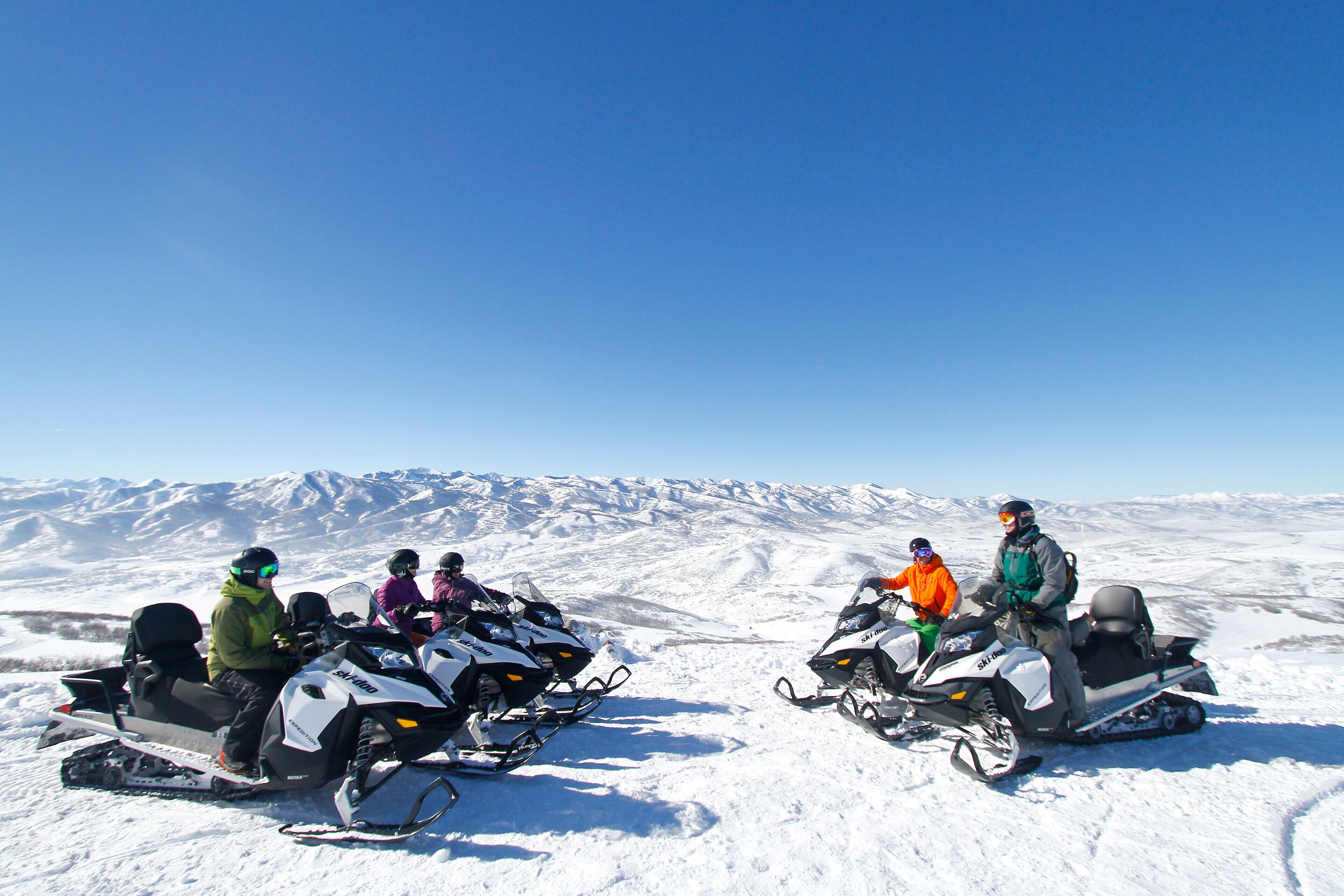 A group of people on snowmobiles on a snowy landscape with mountains in the background.