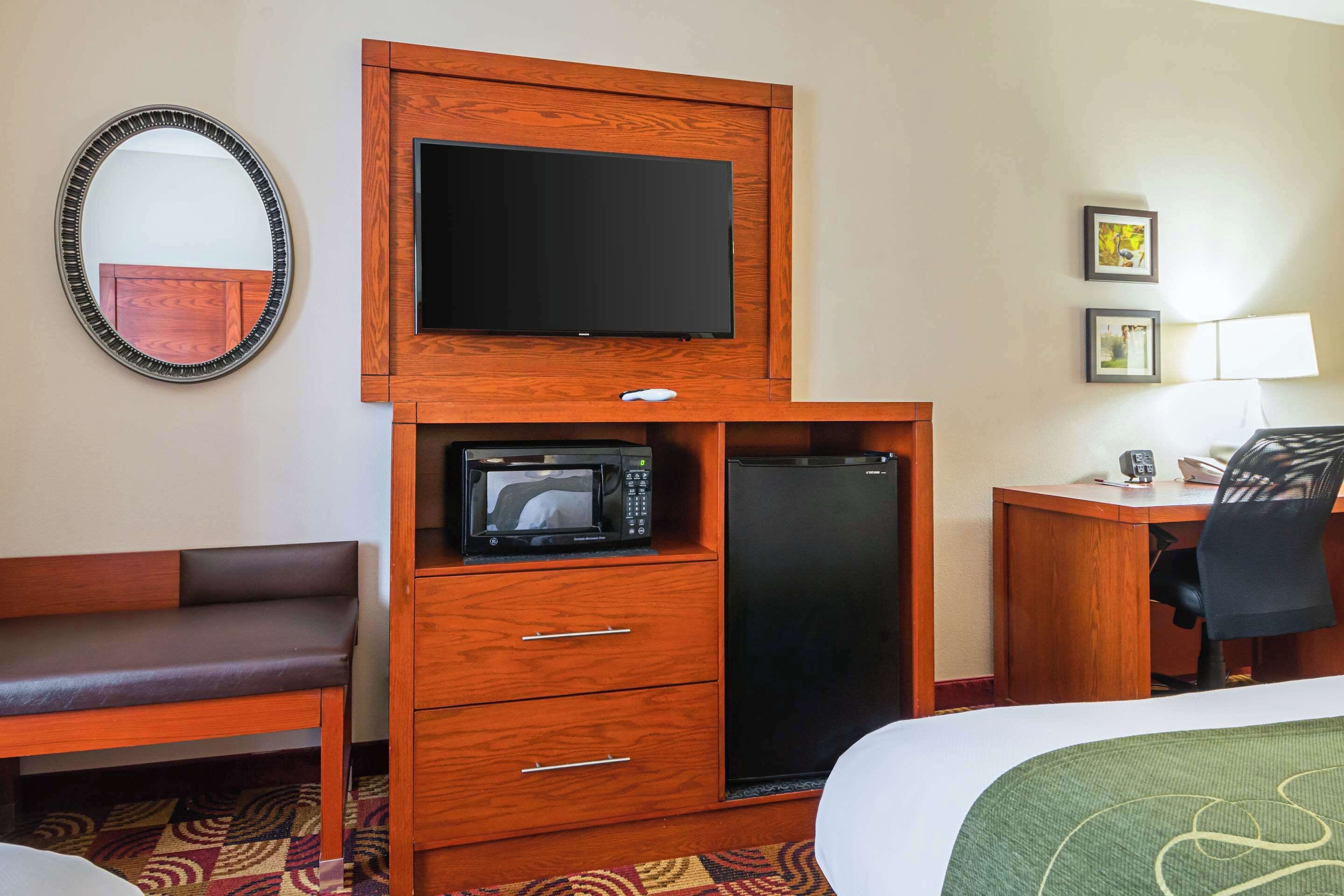 Quarto de hotel com uma secretária de madeira, televisão de tela plana, micro-ondas, frigorífico, cama com colcha verde, cadeira, luminária de mesa e espelho na parede.
