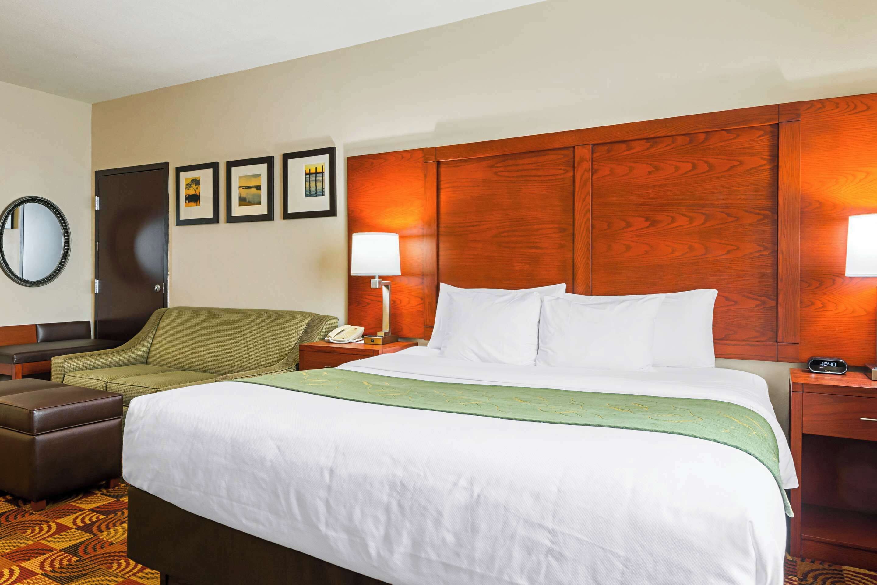 A hotel room with a large bed, a green armchair, a wooden headboard, a nightstand with a lamp, and a wall-mounted mirror.