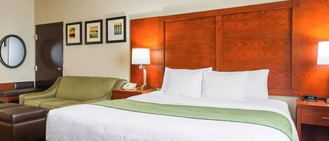 Habitación de hotel con una cama grande, un sillón verde, un cabecero de madera, una mesita de noche con lámpara y un espejo montado en la pared.