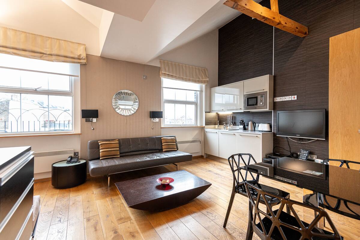 A modern living room with a sofa, coffee table, and dining area. It includes a kitchen with a microwave and a TV.