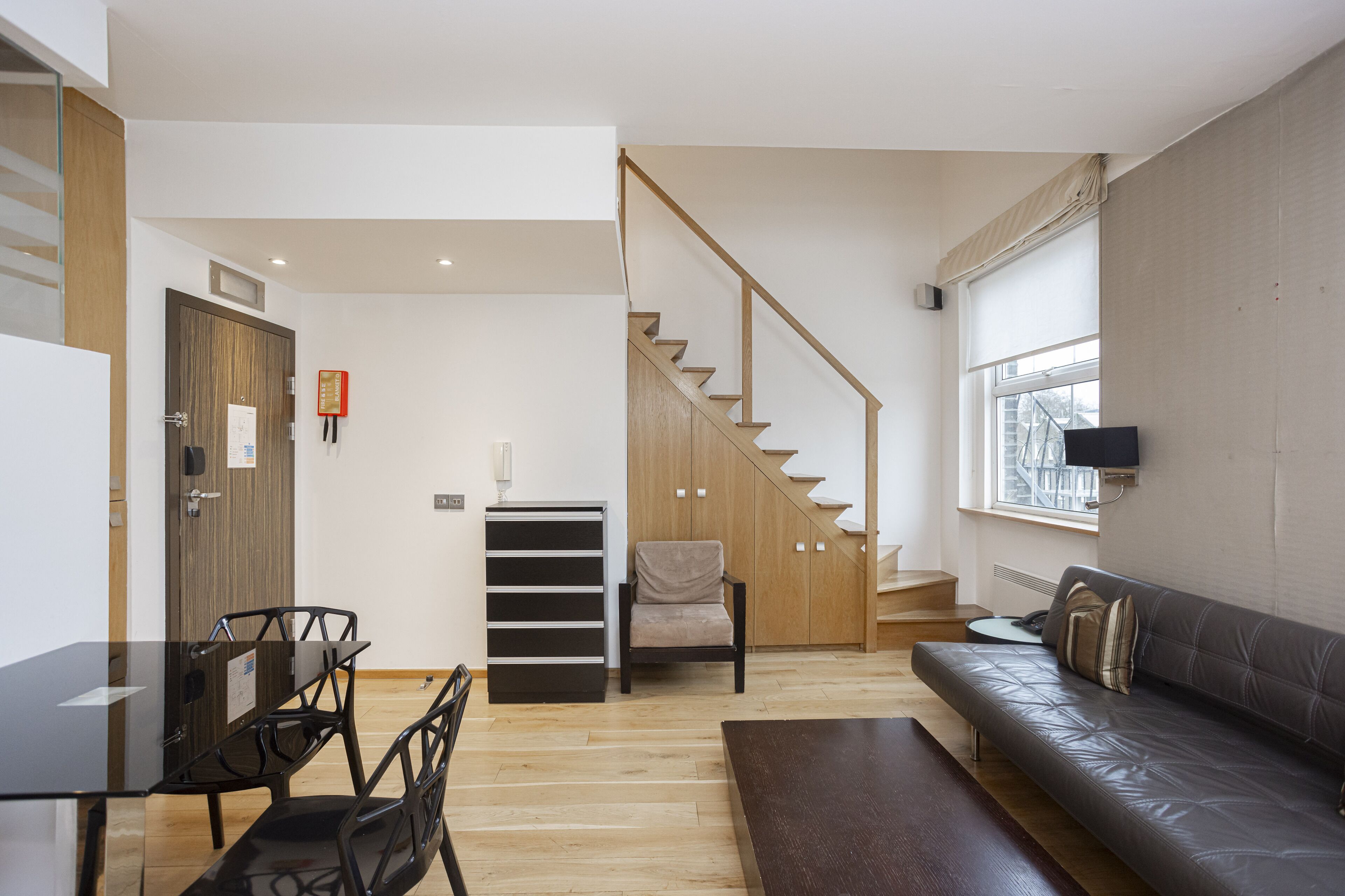 A modern living room with a sofa, a dining table with chairs, a staircase, and a TV mounted on the wall.