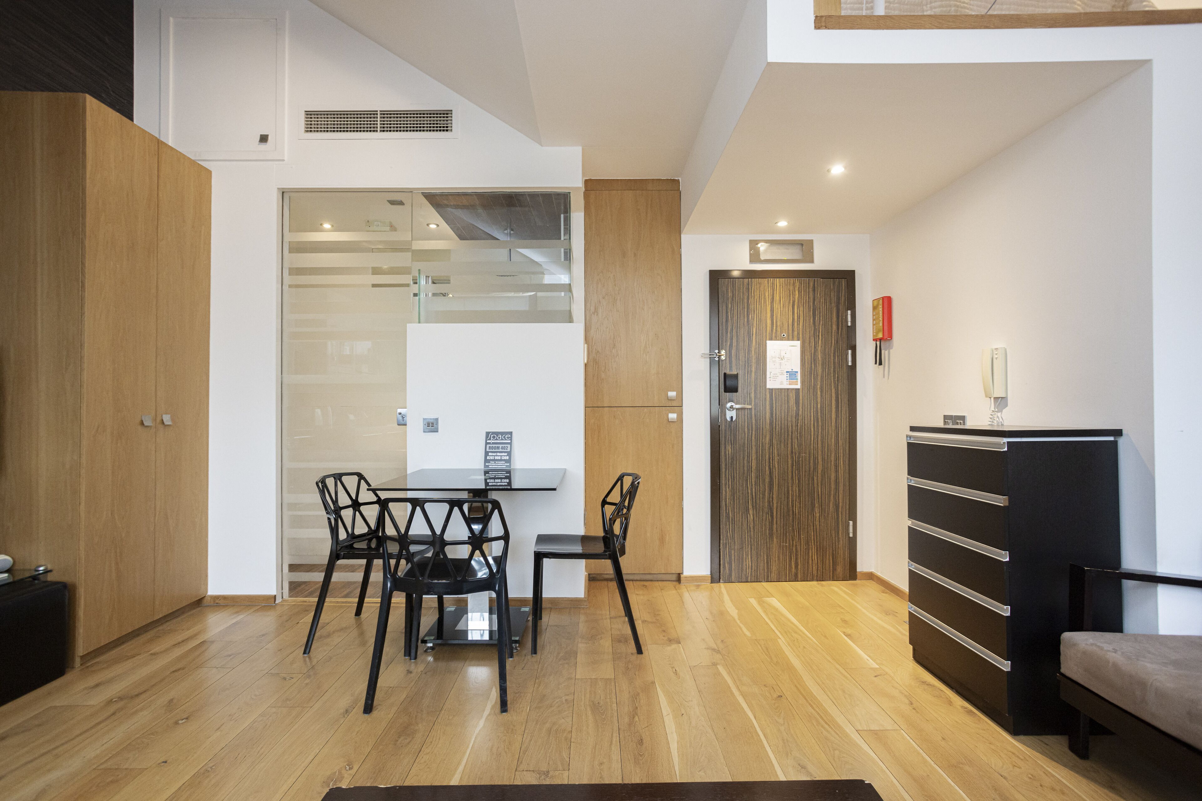 A modern living room with a wooden floor, a glass partition, a dining table with chairs, a sofa, and a cabinet.