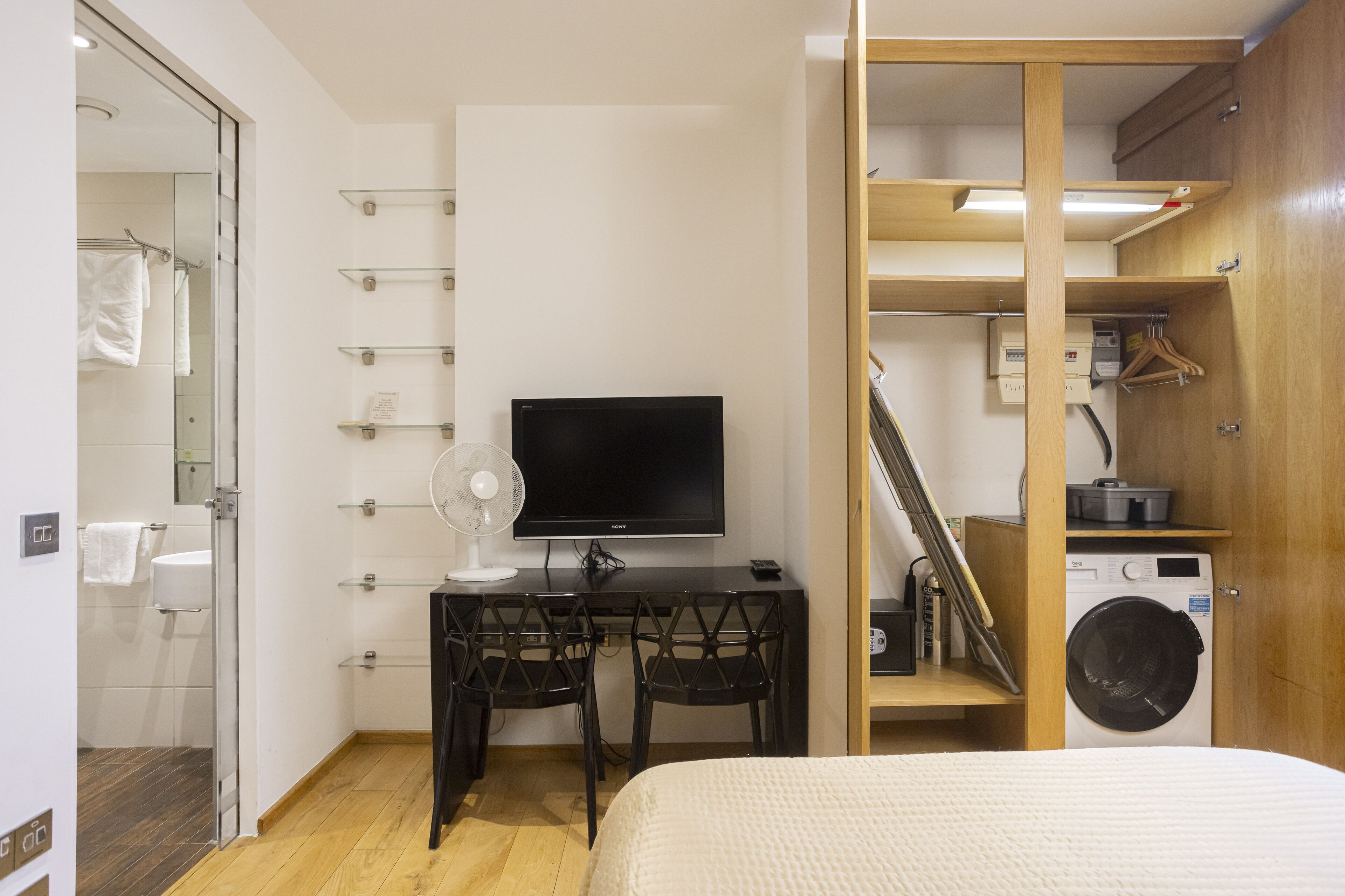 A compact hotel room with a bed, a desk with a TV, a washing machine, and a shower area.
