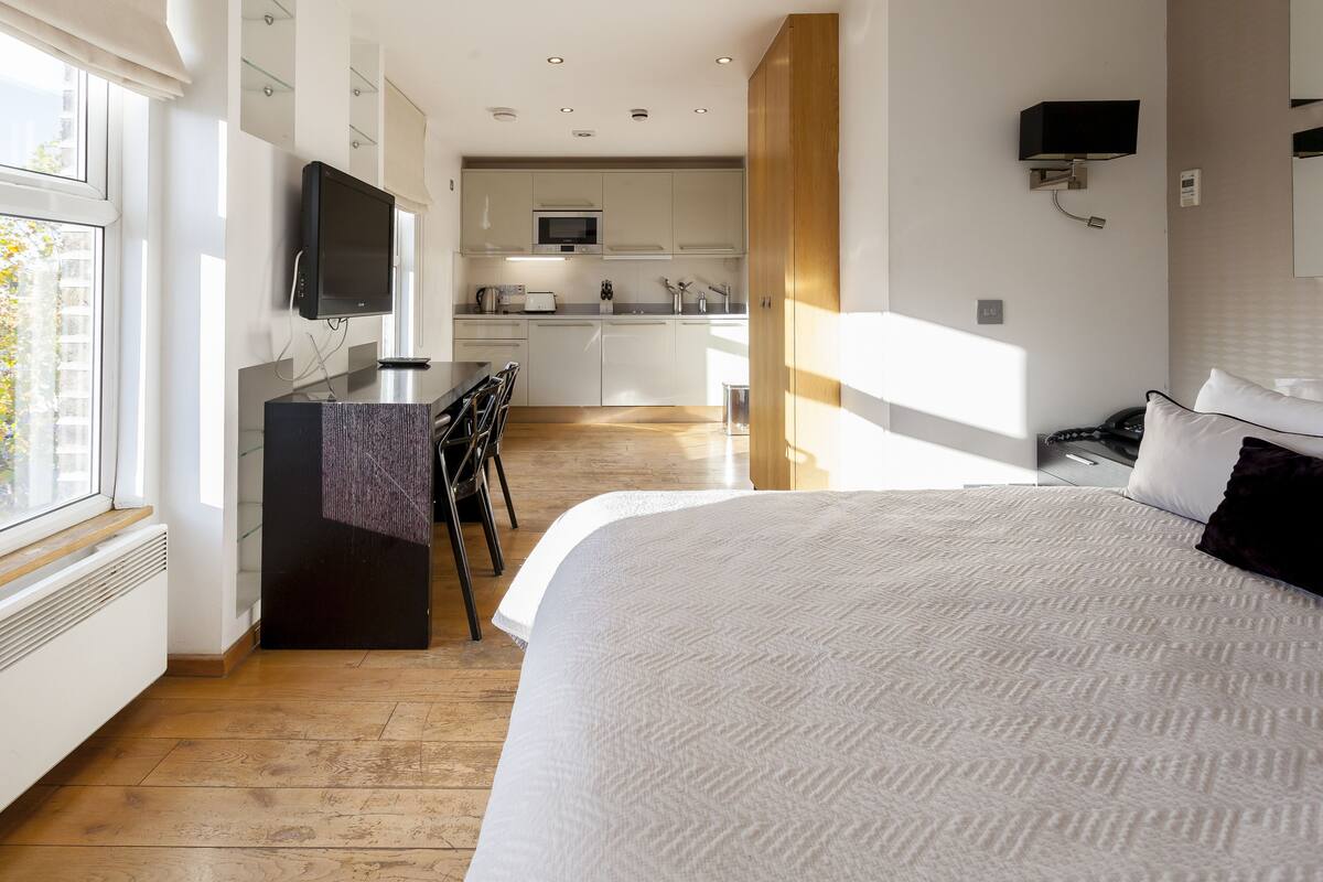 A modern hotel room with a large bed, a desk with a flat-screen TV, a microwave, and a view of the kitchen.