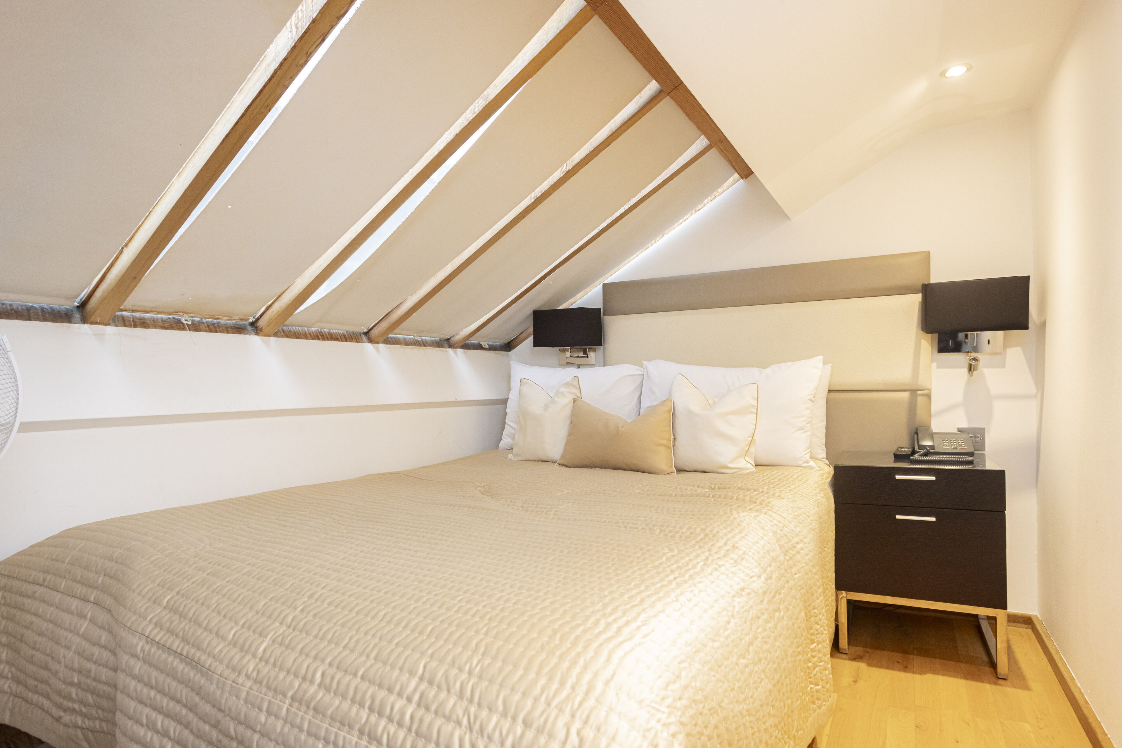 A bedroom with a sloped ceiling, a bed with a beige quilt, a nightstand with a lamp, and a wall-mounted TV.