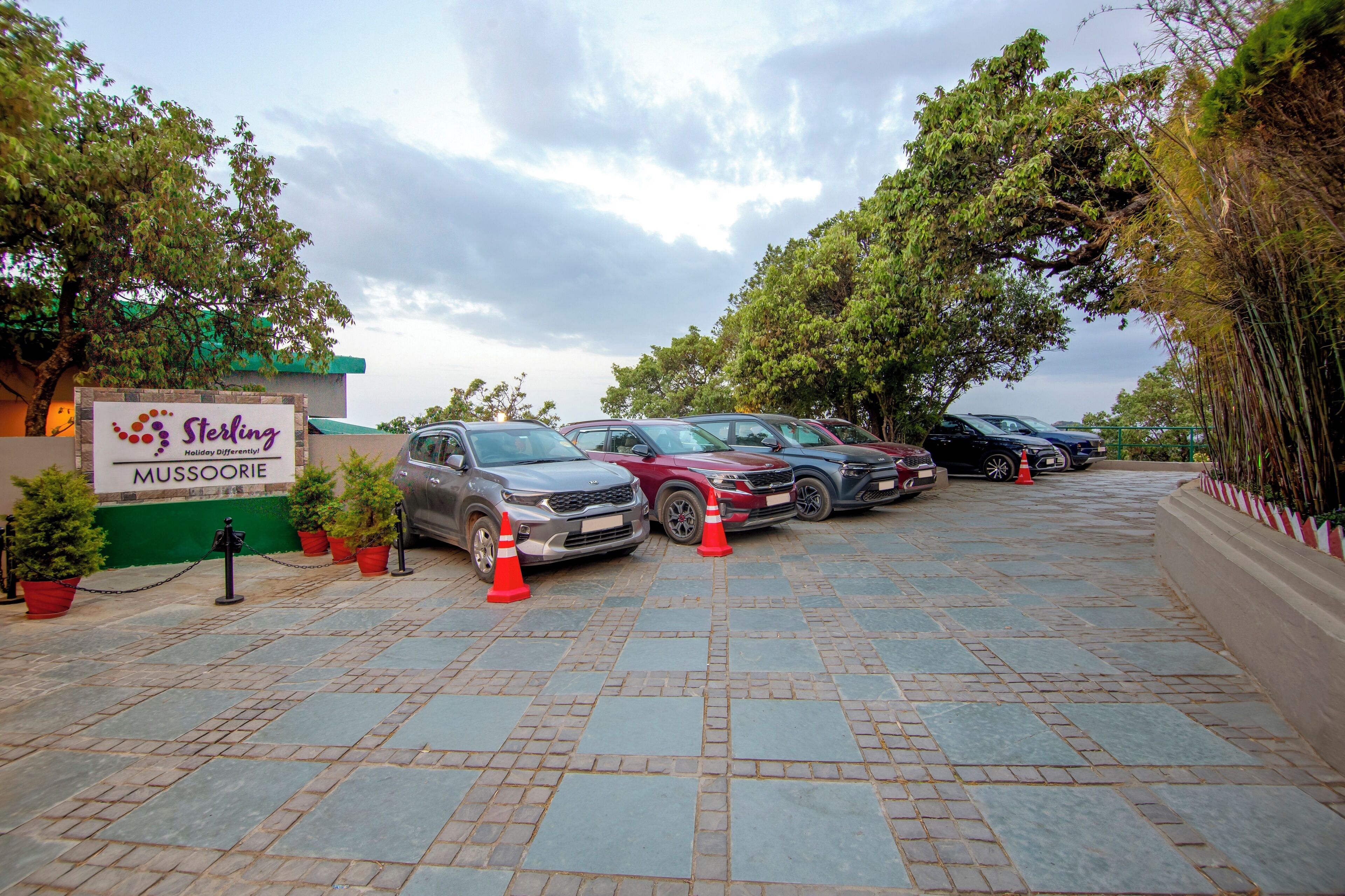 A parking area with several cars, a sign for Sterling Mussoorie, and potted plants along the walkway.