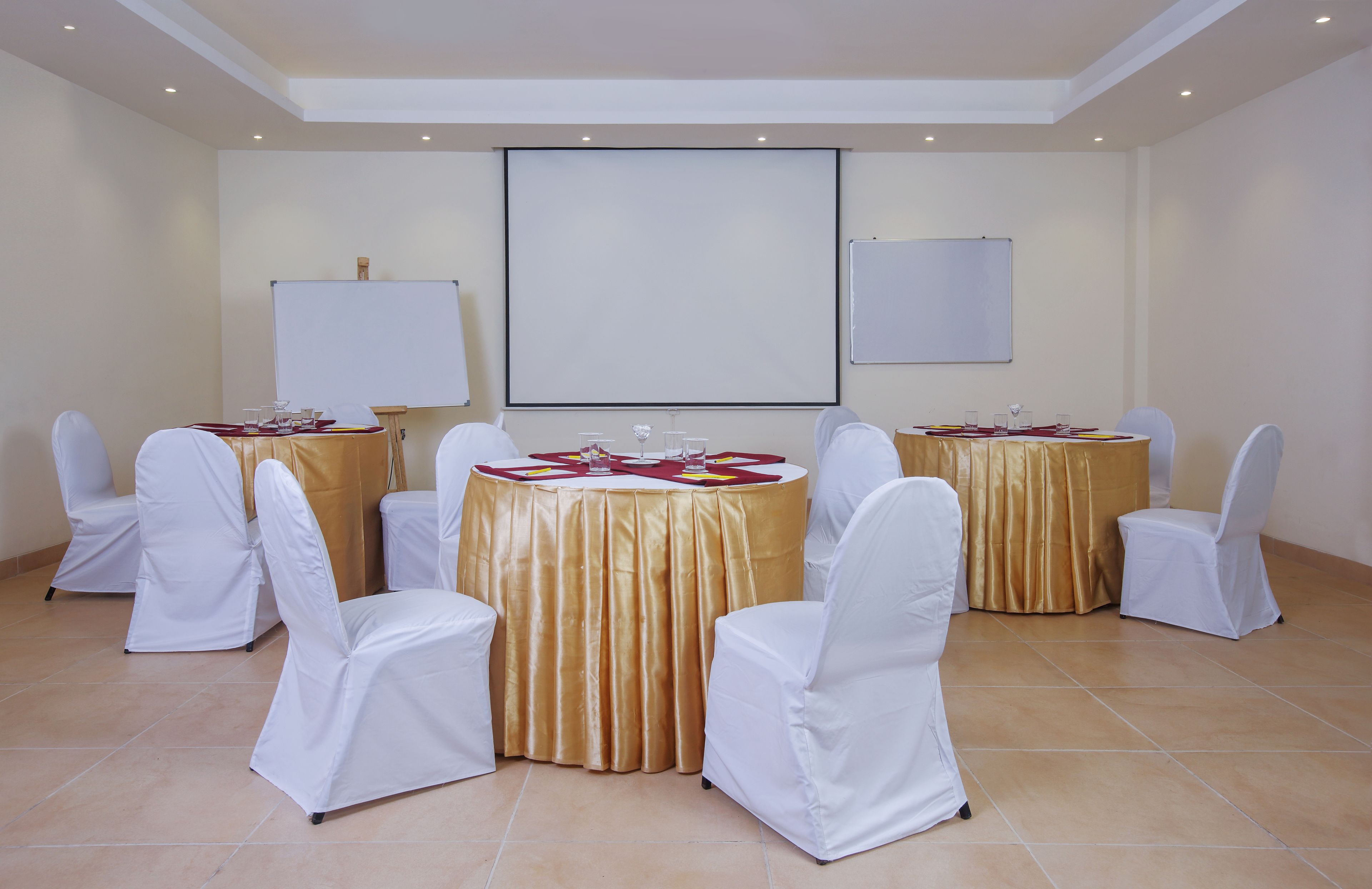 Ein Konferenzraum mit sechs runden Tischen, jeder mit einer goldenen Tischdecke und weißen Stühlen. An den Wänden befinden sich zwei Whiteboards.