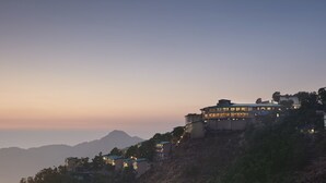 Un edificio de varios pisos con balcones, ubicado en una ladera con montañas al fondo.