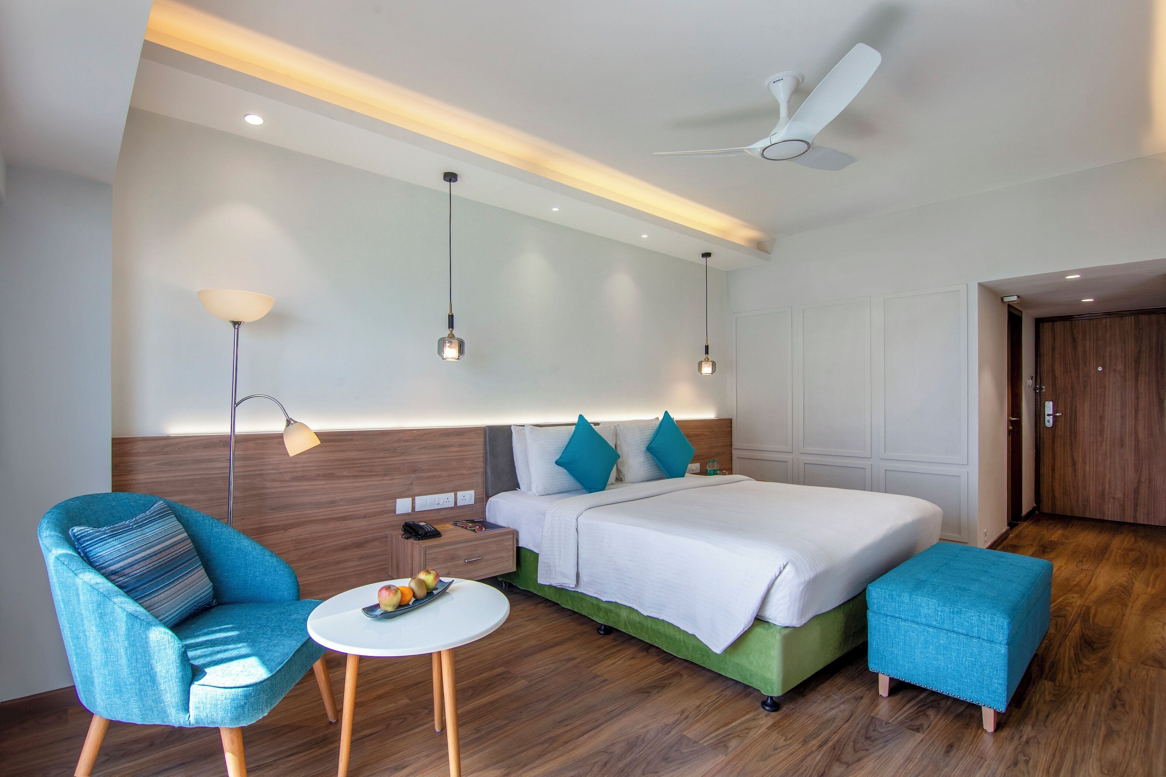A modern hotel room with a bed, a chair, a small table with fruit, and a ceiling fan.