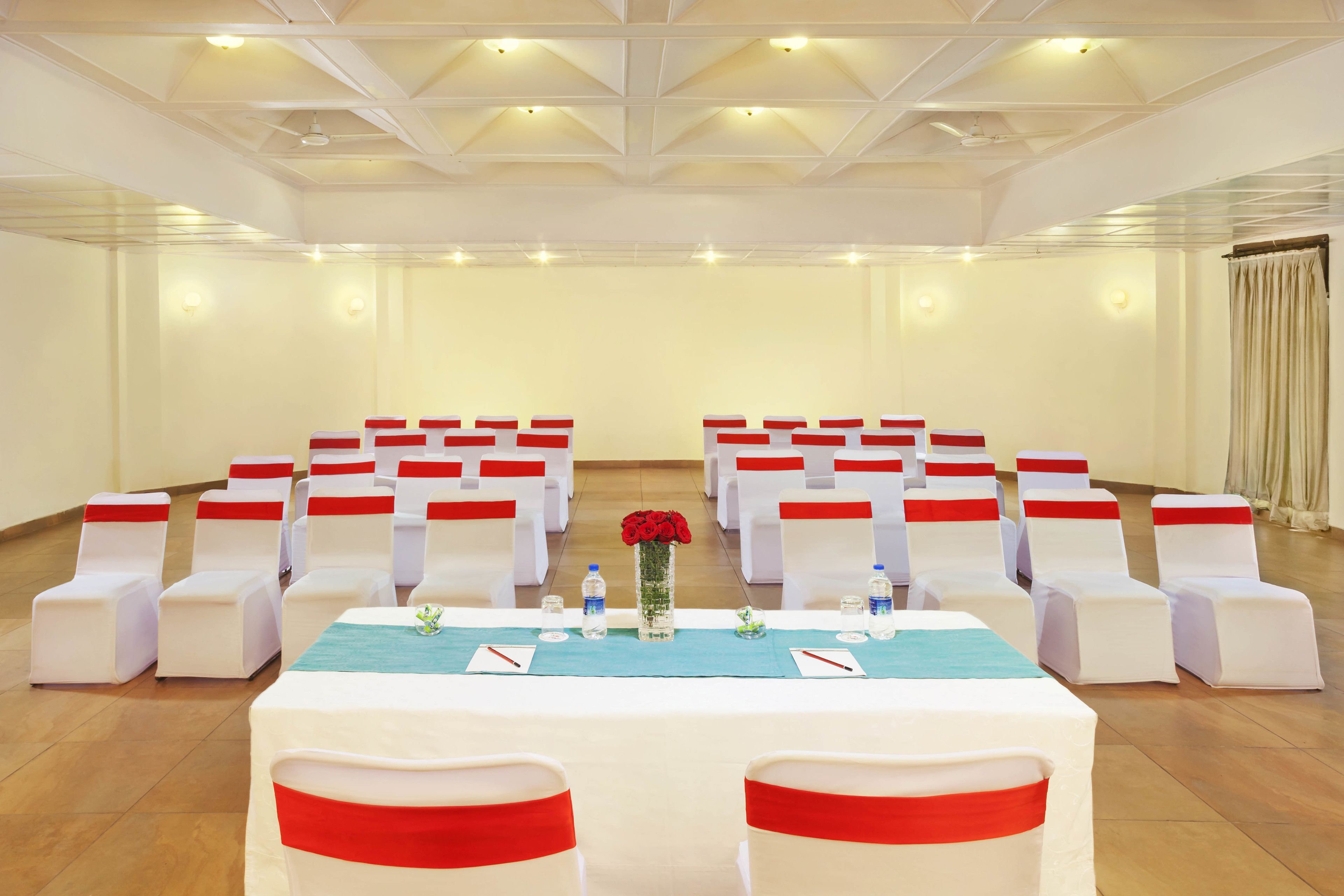 A conference room with rows of white chairs, a long table with a blue tablecloth, and a vase with red flowers.