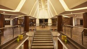 Un vestíbulo de hotel con una escalera central, columnas de madera y un mostrador de recepción.