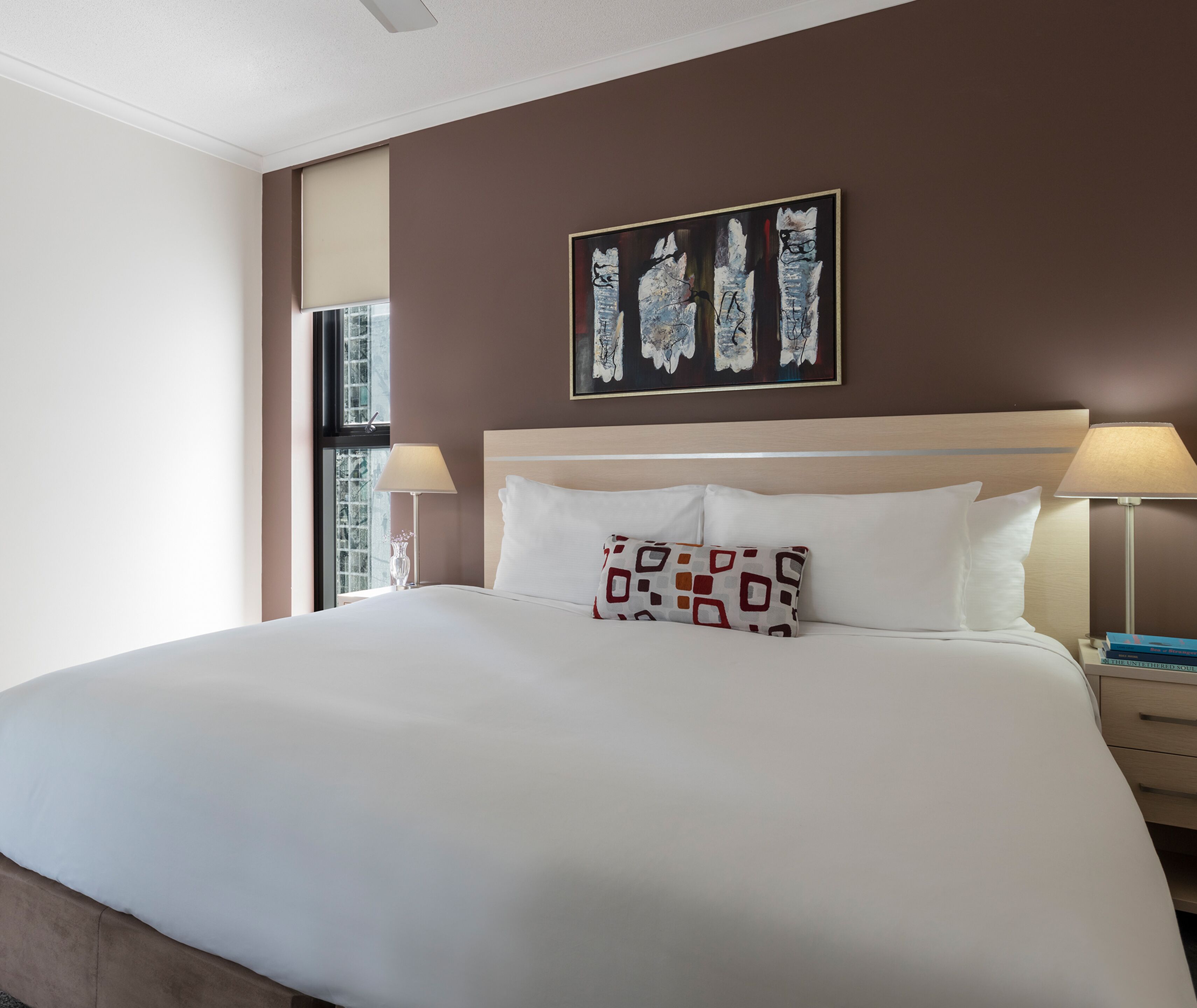Une chambre d’hôtel avec un grand lit, une tête de lit, deux lampes de chevet, une fenêtre et un tableau au mur.