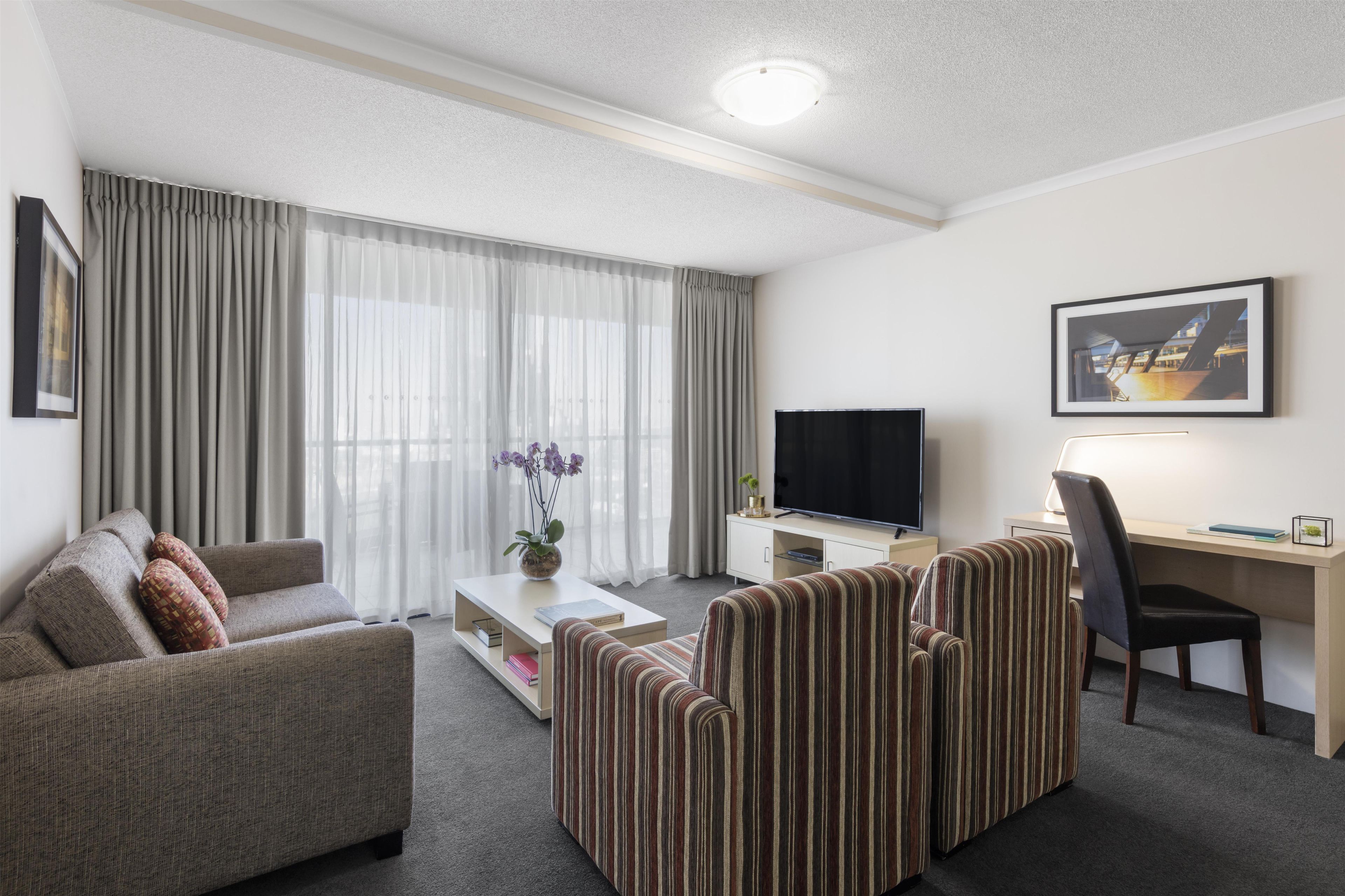 Une chambre d’hôtel avec un canapé, un fauteuil à rayures, un bureau avec un ordinateur, une télévision et un tableau encadré au mur.