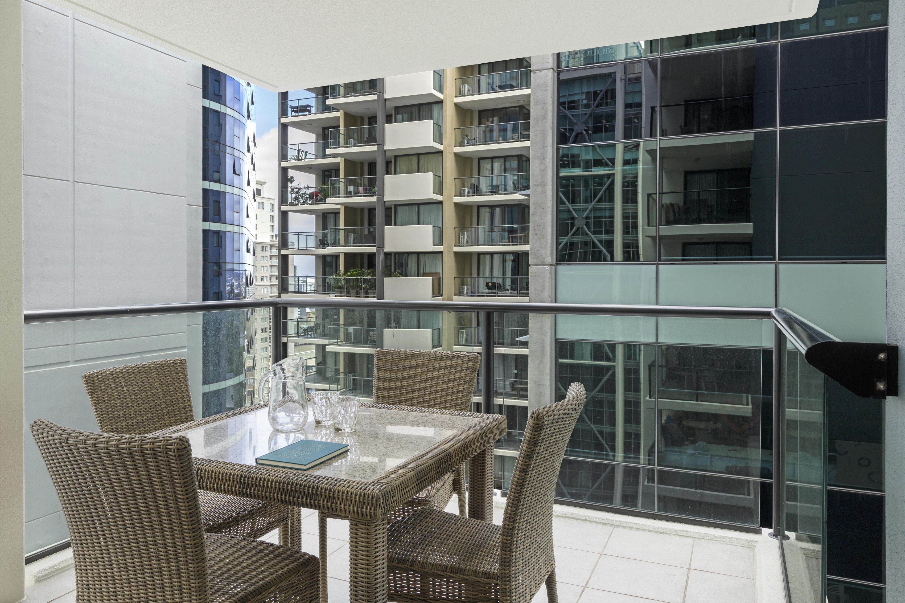 Een balkon met een glazen hekwerk, rieten meubilair en een helder uitzicht op de omliggende gebouwen.