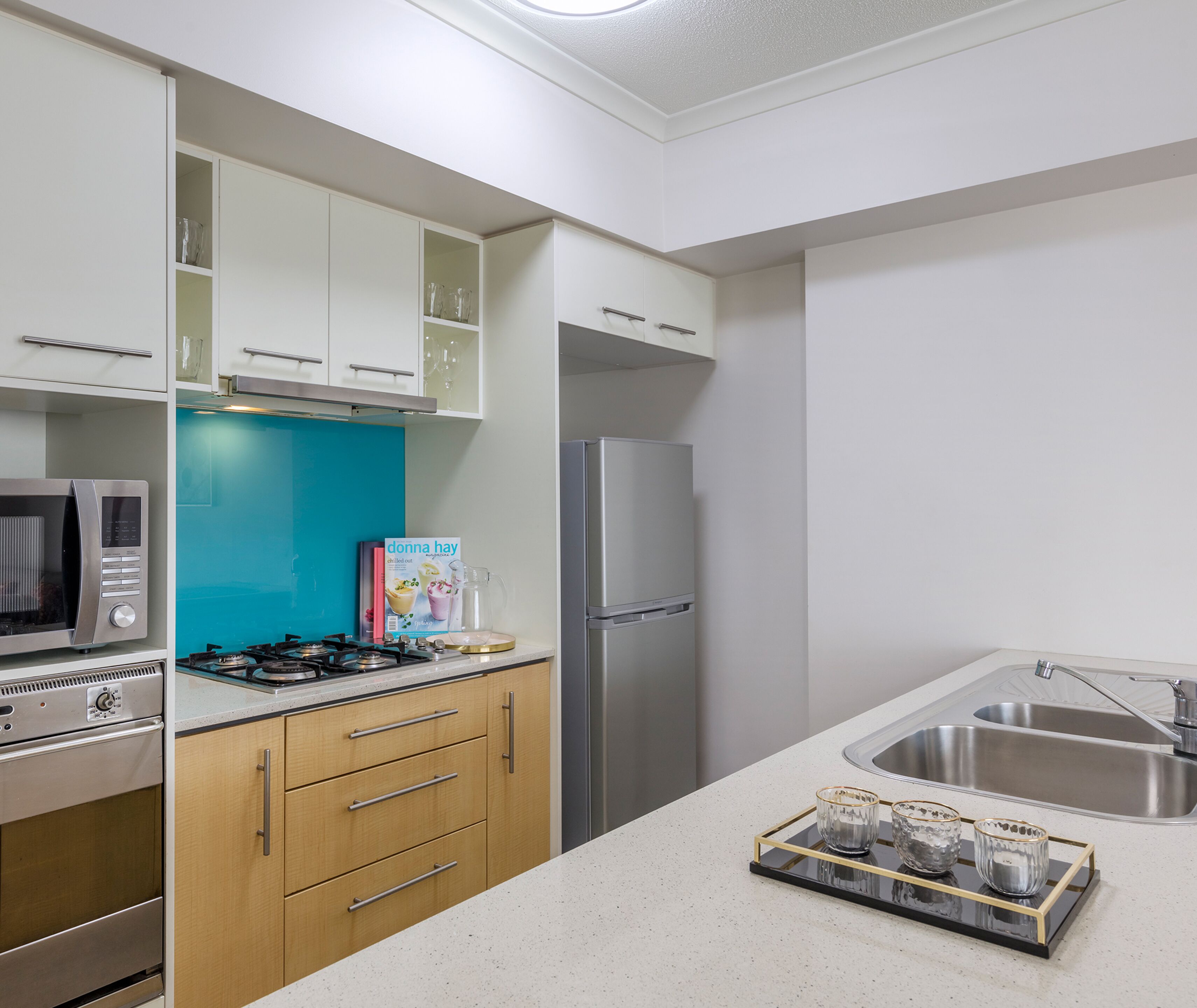 Una cocina moderna con armarios blancos, salpicadero verde azulado, electrodomésticos de acero inoxidable y una mesa de centro de vidrio con tazas.