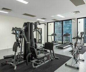 Un gimnasio con diversos equipos de ejercicio, incluyendo cintas de correr y máquinas de pesas, y grandes ventanales con vistas a la ciudad.