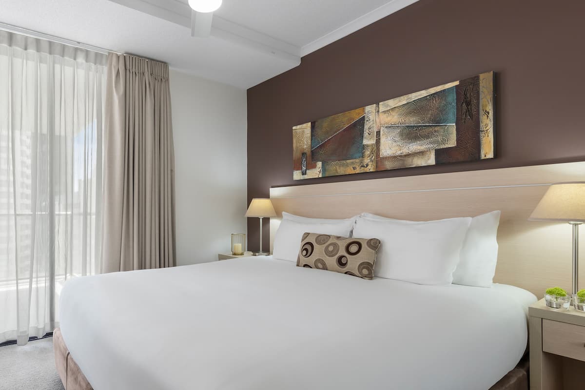 Ein Hotelzimmer mit einem großen Bett, zwei Nachttischen mit Lampen, einer Deckenleuchte und einem Fenster mit Vorhängen.