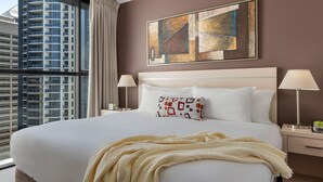Une chambre d’hôtel avec un grand lit, deux lampes de chevet, une fenêtre donnant sur un immeuble de grande hauteur et une œuvre d’art abstraite accrochée au mur.