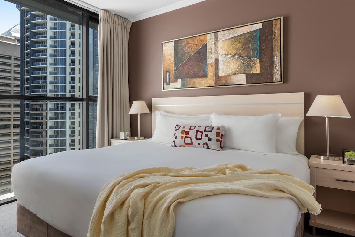 Ein Hotelzimmer mit einem großen Bett, zwei Nachttischlampen, einem Fenster mit Blick auf ein Hochhaus und einem abstrakten Kunstwerk an der Wand.