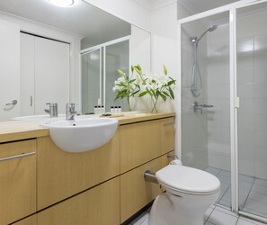 Un baño moderno con un lavamanos blanco, tocador de madera, inodoro y ducha con mampara de cristal.